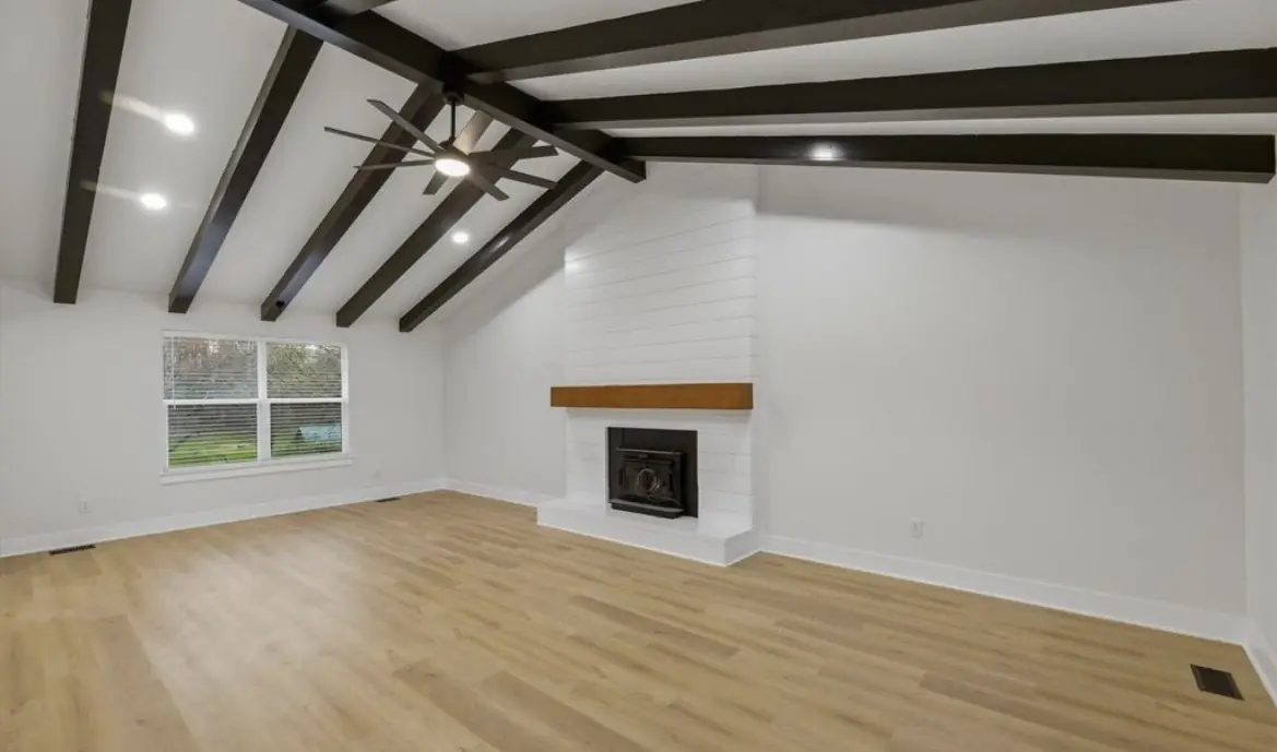 Renovated living room with dark ceiling beams, white walls, a white shiplap fireplace with a wood mantel, recessed lights, and light wood-look plank flooring