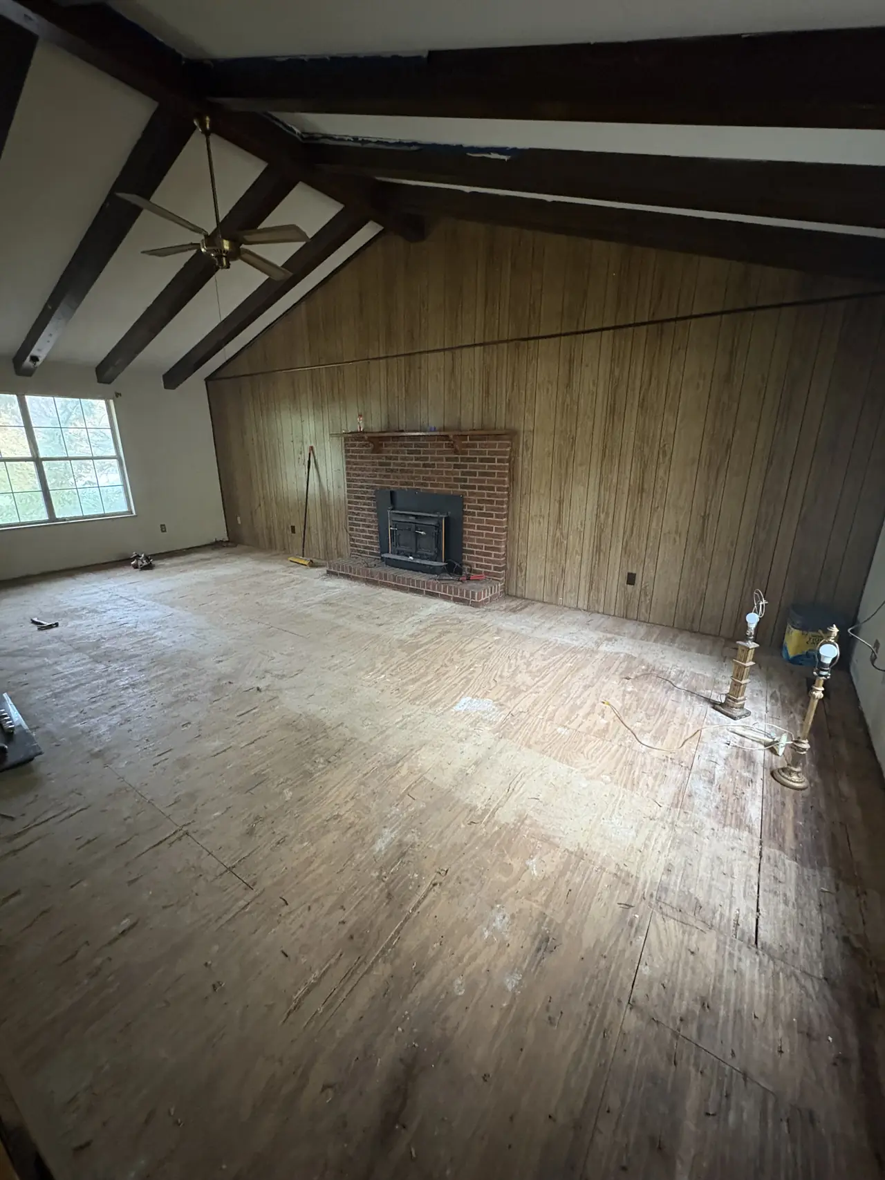 Living room before renovation, with a vaulted beamed ceiling, wood-paneled walls, a brick fireplace, and a bare subfloor
