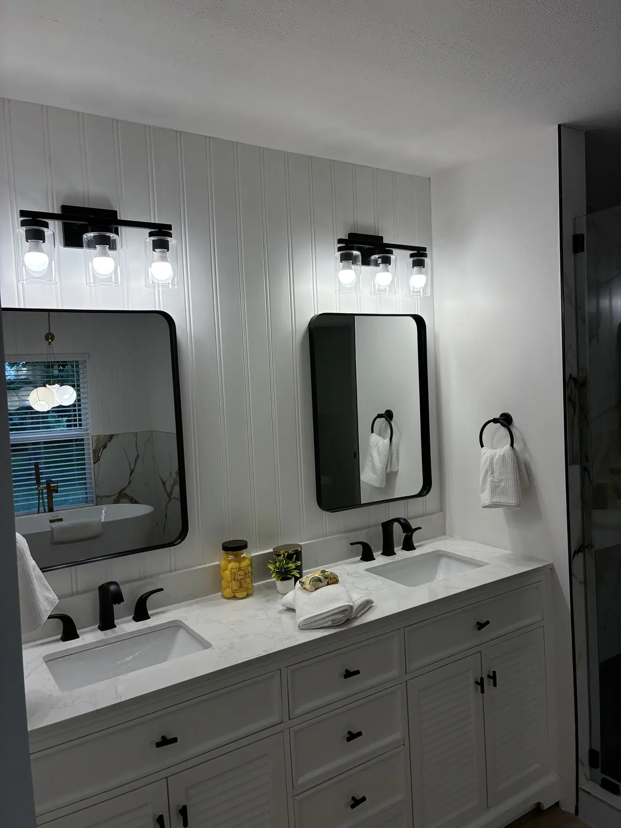 Double vanity with white louvered cabinets, a marble-look countertop, two black-framed mirrors, and a white beadboard wall