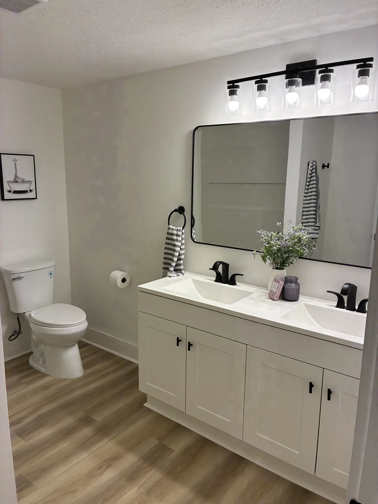 Renovated hall bath with a white double-sink vanity, black faucets and hardware, a black-framed mirror, and wood-look plank flooring