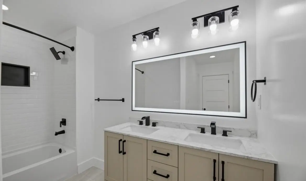 Hall bath with a double vanity in a light wood finish, a lighted mirror, black fixtures, and a tub with a white subway tile surround