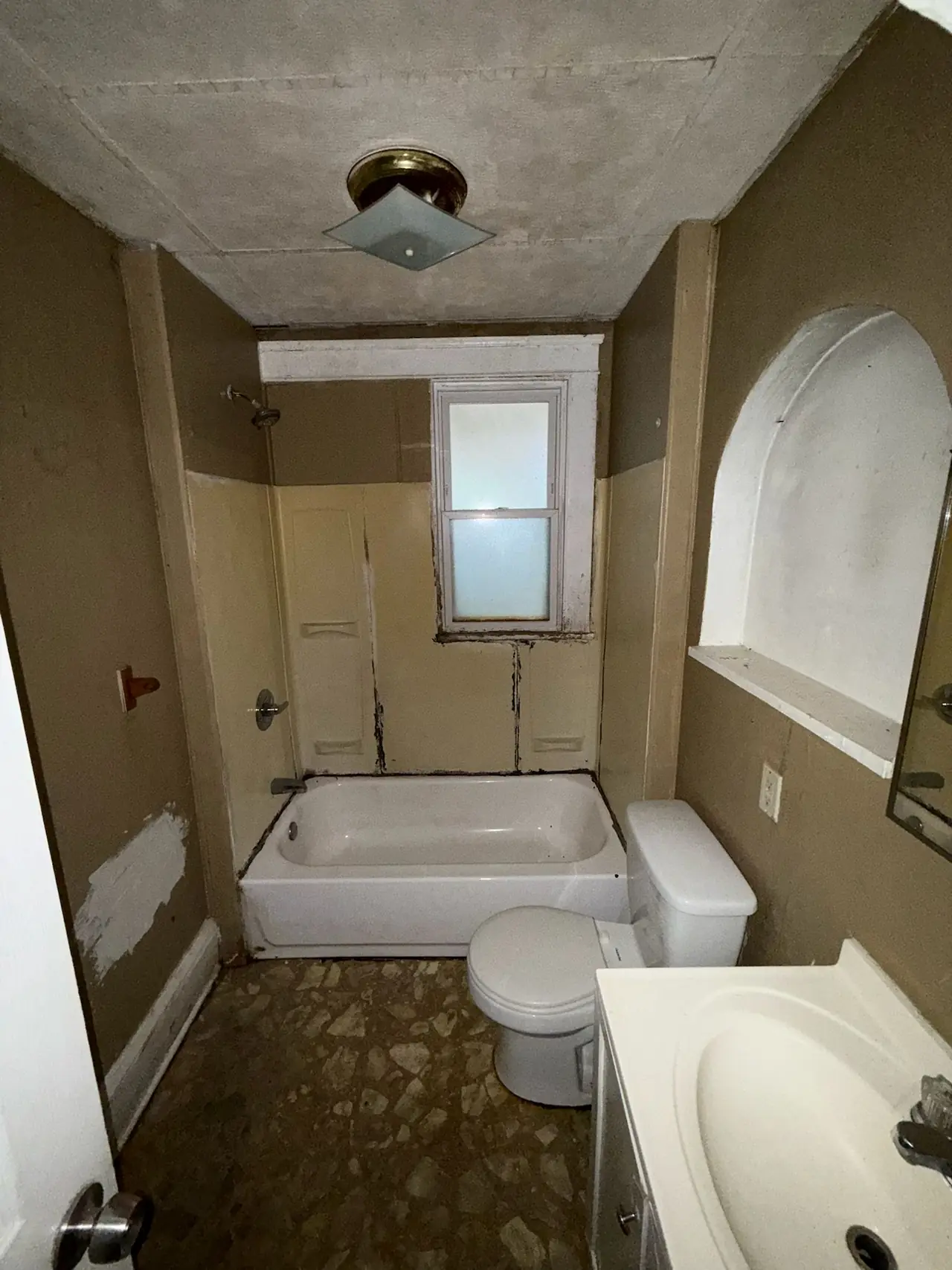 Hall bath before renovation, with an old tub and surround, a toilet and sink, an arched wall niche, and worn flooring