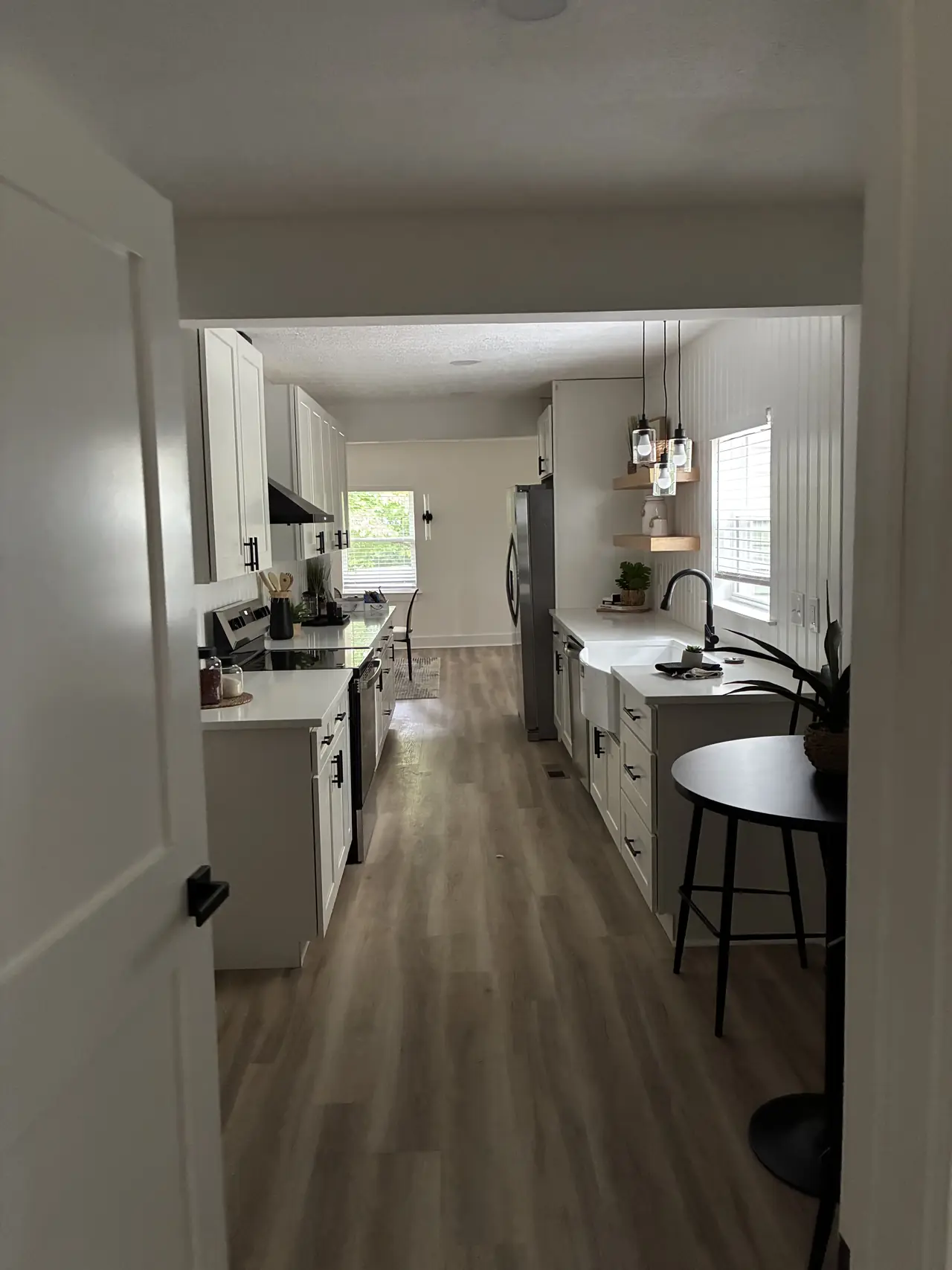 Renovated galley kitchen with white cabinets and countertops, a farmhouse sink, floating wood shelves, glass pendant lights, and wood-look plank flooring