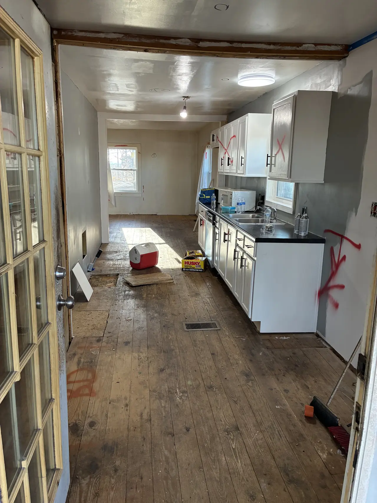 Kitchen before renovation: worn wood plank floor, old white cabinets with a black countertop, and red demolition marks on the walls