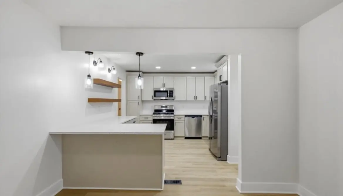 Renovated kitchen seen from the dining area: a peninsula countertop, glass pendant lights, floating wood shelves, white shaker cabinets, and stainless appliances
