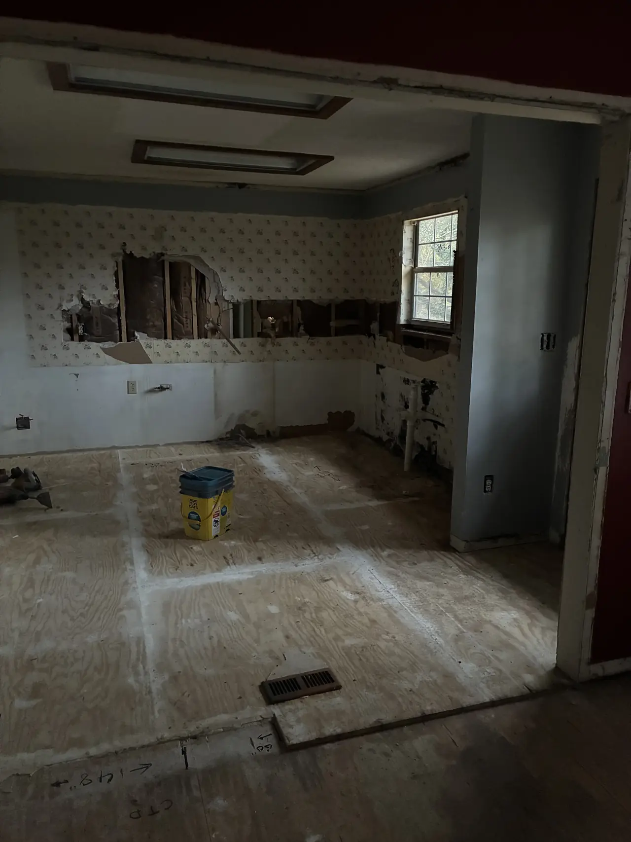 Kitchen during demolition, with the old cabinets removed, wallpaper torn open, and a bare plywood subfloor
