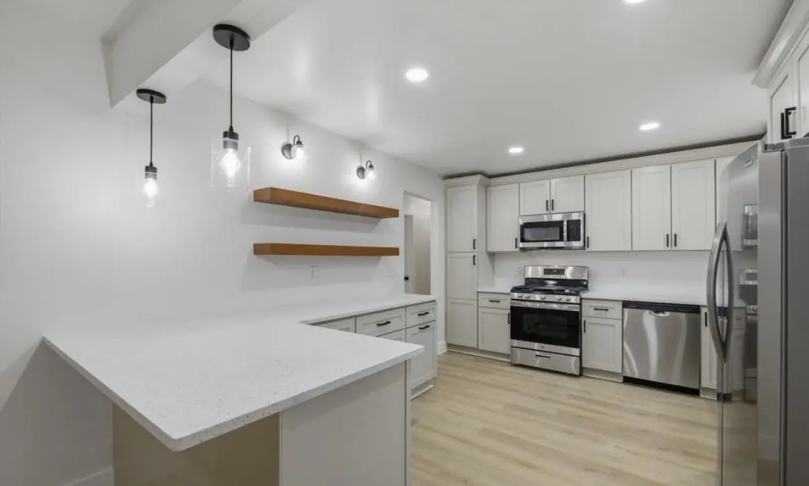 Kitchen with a white peninsula countertop, glass pendant lights, floating wood shelves, white shaker cabinets, a gas range, and stainless appliances