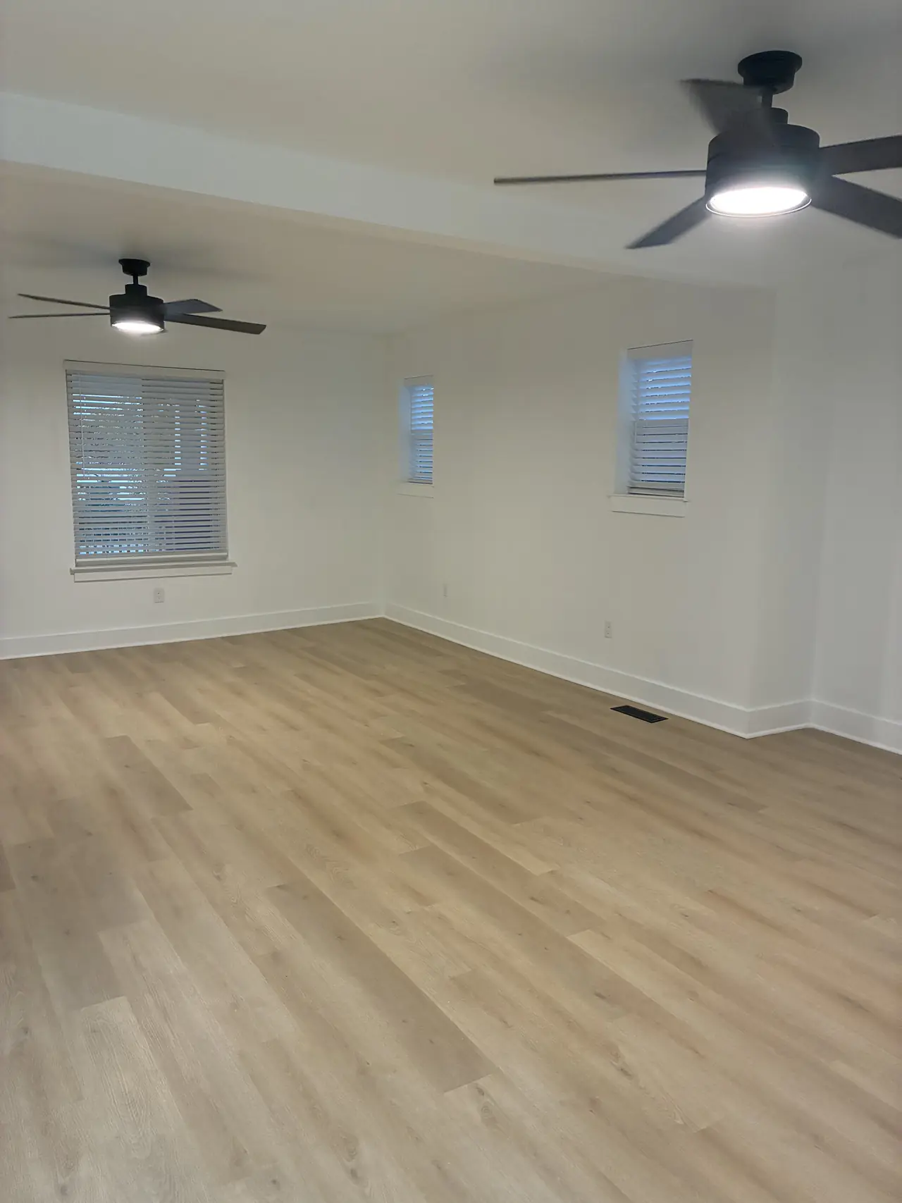 Living room after renovation, opened into one large room with white walls, two black ceiling fans, new blinds, and light wood-look plank flooring