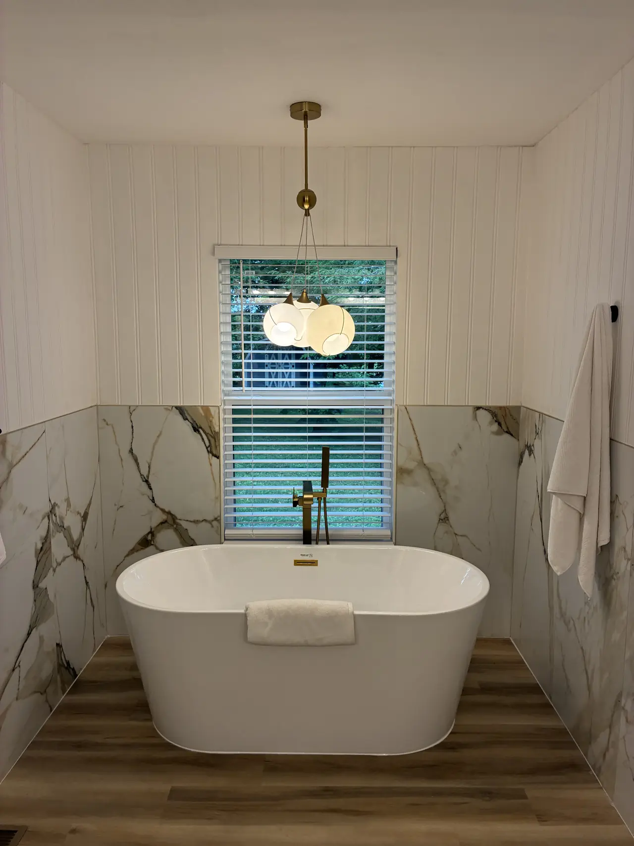 Renovated primary bath with a freestanding soaker tub, marble-look tile, and a brass floor-mounted tub filler