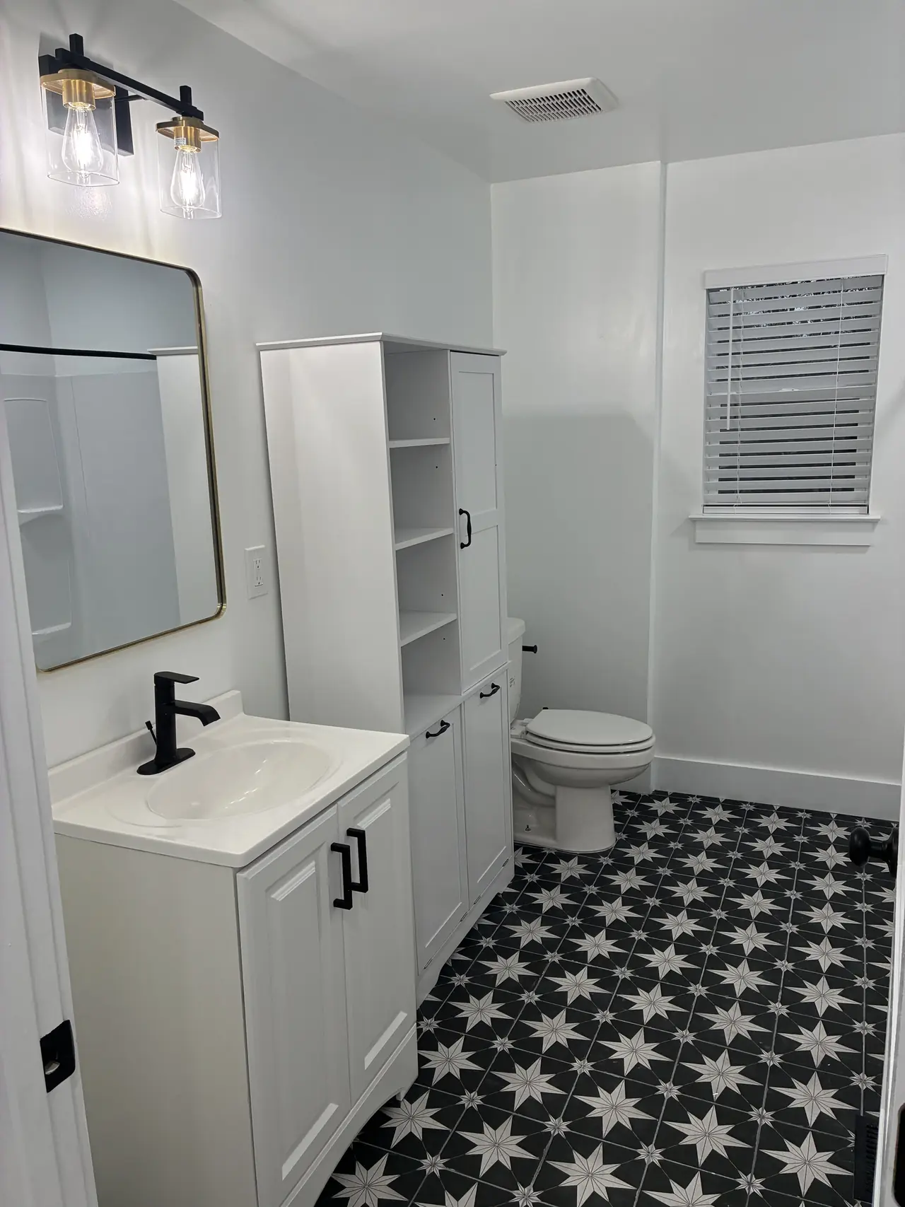 Bath with a white vanity, a tall white storage cabinet, a black-and-white star-pattern tile floor, and black fixtures